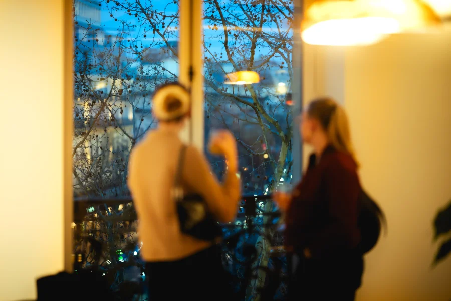 Silhouette floue et artistique de deux invitées discutant près d'une fenêtre lors d'une soirée corporate à Paris, vue sur les arbres illuminés en basse lumière.
